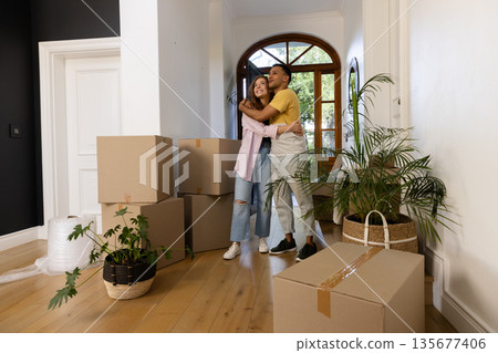 Young couple embracing in new home surrounded by moving boxes and plants Young couple embracing in new home surrounded by moving boxes and plants 135677406