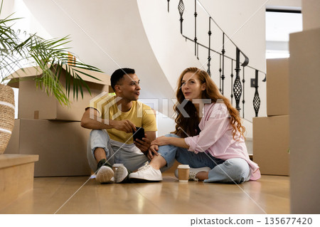 Young couple sitting on floor surrounded by moving boxes, enjoying coffee together 135677420