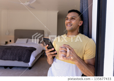 Smiling man holding smartphone and coffee mug, relaxing in bedroom doorway 135677433