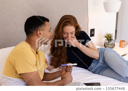 Young couple on bed holding pregnancy test, expressing surprise and joy together Young couple on bed holding pregnancy test, expressing surprise and joy together 135677448
