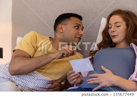 Young couple at home, excitedly looking at ultrasound photo together on bed Young couple at home, excitedly looking at ultrasound photo together on bed 135677449