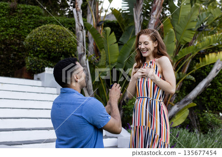 Young man proposing to surprised woman in garden, both smiling with joy 135677454