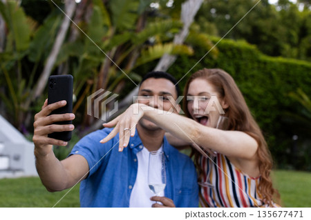 Young couple taking selfie outdoors, woman showing engagement ring excitedly 135677471