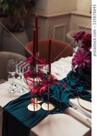 Luxurious banquet table setting with beige tablecloth, green napkins in gold holders, red candles in golden candlesticks and ceramic plates. Refined festive dining decor. Luxurious banquet table setting with beige tablecloth, green napkins in gold holders, red candles in golden candlesticks and ceramic plates. Refined festive dining decor. 135678043
