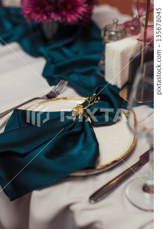 Luxurious banquet table setting with beige tablecloth, green napkins in gold holders, red candles in golden candlesticks and ceramic plates. Refined festive dining decor. Luxurious banquet table setting with beige tablecloth, green napkins in gold holders, red candles in golden candlesticks and ceramic plates. Refined festive dining decor. 135678045