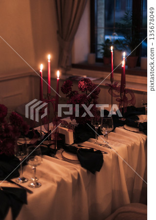 Luxurious banquet table setting with beige tablecloth, green napkins in gold holders, red candles in golden candlesticks and ceramic plates. Refined festive dining decor. 135678049