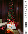 Luxurious Christmas tree decorated with lights and ornaments in a cozy interior. White gift bags under the tree, blurred tangerines in foreground creating festive holiday mood. 135678193