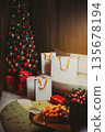 Luxurious Christmas tree decorated with lights and ornaments in a cozy interior. White gift bags under the tree, blurred tangerines in foreground creating festive holiday mood. 135678194