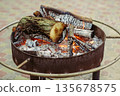 Photo of a round outdoor fire pit used for warming hands in winter. Burning firewood and glowing coals create a cozy atmosphere in an open space. 135678575