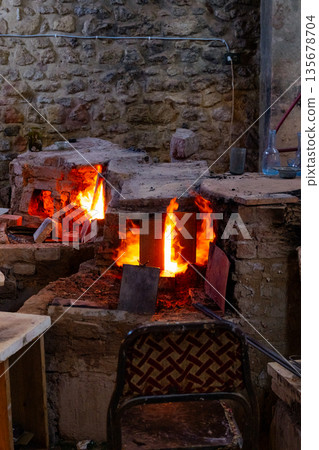 : A traditional glass furnace with bright flames and glowing embers. The structure is made of stone and metal, showcasing the glass-making process. 135678704