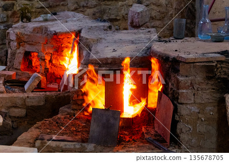 : A traditional glass furnace with bright flames and glowing embers. The structure is made of stone and metal, showcasing the glass-making process. 135678705