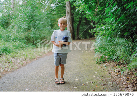 Young child walking along a forest path holding a piece of clothing. The child stands confidently on the path surrounded by lush greenery, conveying a sense of adventure and freedom. Young child walking along a forest path holding a piece of clothing. The child stands confidently on the path surrounded by lush greenery, conveying a sense of adventure and freedom. 135679076