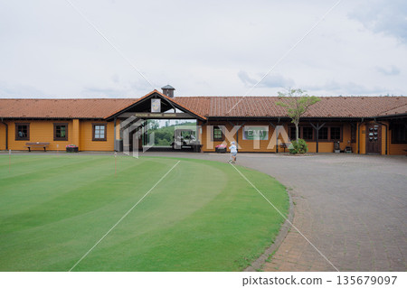 Scenic view of a golf clubhouse with wooden architecture and a child joyfully riding a scooter along the path. The setting exudes leisure and outdoor enjoyment amidst nature. Scenic view of a golf clubhouse with wooden architecture and a child joyfully riding a scooter along the path. The setting exudes leisure and outdoor enjoyment amidst nature. 135679097