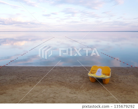 Yellow pedal boat on sandy beach next to calm lake water with reflection of sky. Peaceful waterscape for vacation travel. Yellow pedal boat on sandy beach next to calm lake water with reflection of sky. Peaceful waterscape for vacation travel. 135679292