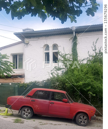Red car parked on a street next to a white house with green foliage. Classic vehicle outdoors for urban setting. 135679296