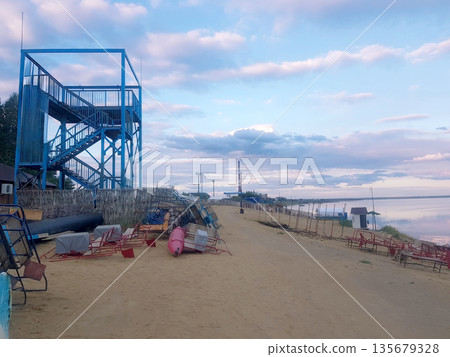 Blue observation tower on a sandy beach by a lake with abandoned paddle boats. Calm evening landscape with industrial background. Blue observation tower on a sandy beach by a lake with abandoned paddle boats. Calm evening landscape with industrial background. 135679328