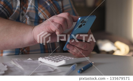 A person interacts with a smartphone using both hands while sitting at a table filled with papers and a keyboard. The light from a window brightens the area. A person interacts with a smartphone using both hands while sitting at a table filled with papers and a keyboard. The light from a window brightens the area. 135679571