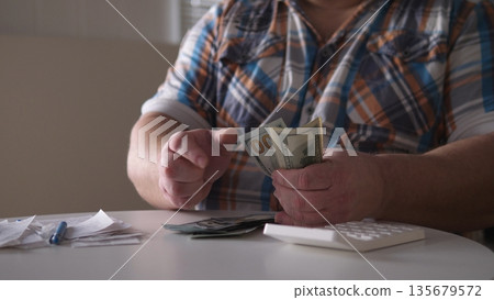 A person sits at a table counting bills and looking at various receipts in a bright room. A calculator is present for calculations while handling cash and documents. A person sits at a table counting bills and looking at various receipts in a bright room. A calculator is present for calculations while handling cash and documents. 135679572