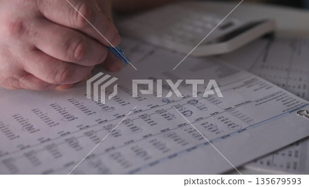 A person uses a pen to mark details on printed documents while a calculator sits nearby. The scene shows an office setup focused on financial tasks and planning. A person uses a pen to mark details on printed documents while a calculator sits nearby. The scene shows an office setup focused on financial tasks and planning. 135679593