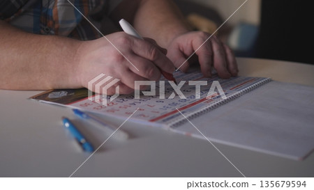 A person focuses on writing numbers on a calendar with a red pen. Two blue pens are placed on the table. The scene takes place indoors during the evening hours. A person focuses on writing numbers on a calendar with a red pen. Two blue pens are placed on the table. The scene takes place indoors during the evening hours. 135679594