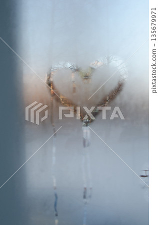 A heart-shaped clear patch melted into frosty glass, glowing warmly against cold blue condensation. 135679971