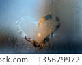 A heart-shaped clear patch melted into frosty glass, glowing warmly against cold blue condensation. 135679972