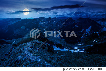 path through mountain ridge with snow on distant green hills at night. gorgeous panoramic view of highland scenery for recreation in summer in full moon light. inner strength to overcome fear 135680008