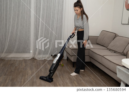 Woman Cleans Living Room Floor With Vacuum While Pet Sits Nearby in Cozy Home Space Woman Cleans Living Room Floor With Vacuum While Pet Sits Nearby in Cozy Home Space 135680594