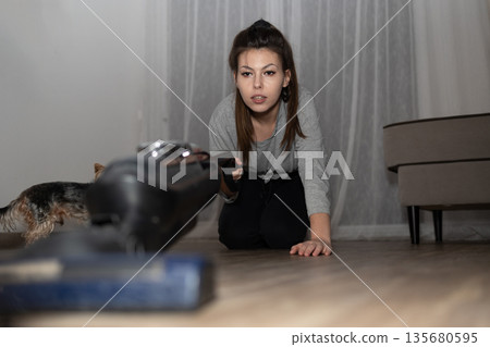 Young Person Cleans Floor With Vacuum While Looking Focused in Living Room 135680595
