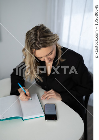 Woman in Black Jacket Writing in Notebook on Table Indoors 135680649