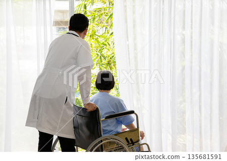 Doctor standing behind an elderly woman in a wheelchair, both looking out the window. The scene captures a calm, caring moment, symbolizing hope, recovery, and compassionate elderly care 135681591