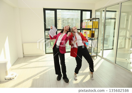 Two joyful women celebrating success in a modern office space, throwing papers in the air during a bright afternoon 135682586