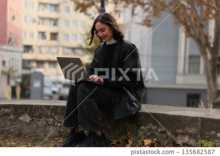 Focused young woman works on laptop in urban park, surrounded by fall foliage and city buildings on a cool day Focused young woman works on laptop in urban park, surrounded by fall foliage and city buildings on a cool day 135682587