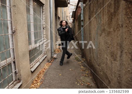Young person joyfully walking through a narrow alley surrounded by weathered buildings and fallen leaves in autumn 135682599