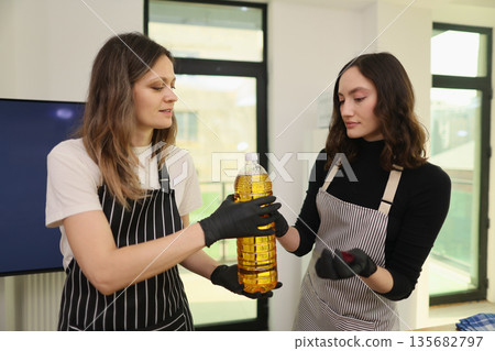 Two women engaged in a cooking lesson, sharing tips with a bottle of cooking oil in a bright kitchen setting 135682797
