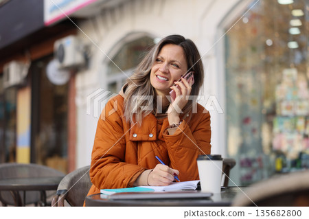 Smiling woman talks on the phone while seated outdoors in a cozy cafe during a sunny day, enjoying her warm coffee Smiling woman talks on the phone while seated outdoors in a cozy cafe during a sunny day, enjoying her warm coffee 135682800