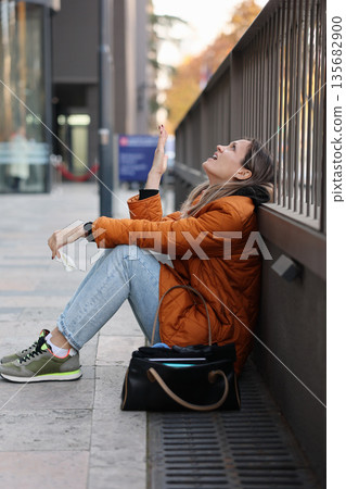 Woman enjoying a moment of laughter while sitting on a city sidewalk in autumn Woman enjoying a moment of laughter while sitting on a city sidewalk in autumn 135682900