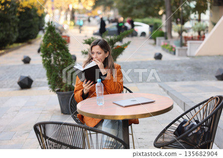 Woman enjoys a quiet moment reading a book at a lively outdoor caf during autumn afternoons Woman enjoys a quiet moment reading a book at a lively outdoor caf during autumn afternoons 135682904
