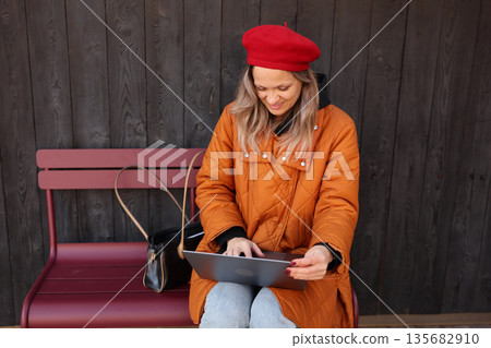 Young woman in a red beret enjoys her time on a laptop while sitting on a bench outside a cozy wooden building Young woman in a red beret enjoys her time on a laptop while sitting on a bench outside a cozy wooden building 135682910