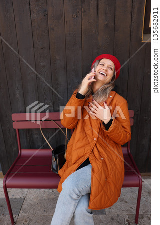 Joyful woman chats on her phone while sitting on a bench against a rustic wooden wall in a cozy urban setting 135682911