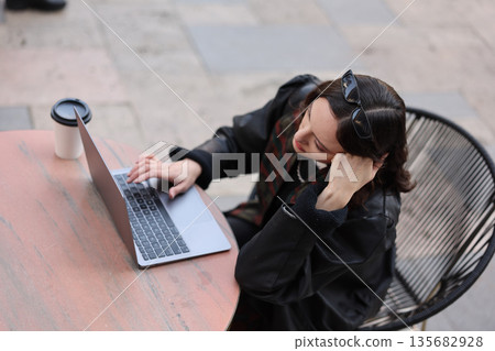 Young woman working on laptop outside while enjoying coffee in a lively urban setting during daylight hours Young woman working on laptop outside while enjoying coffee in a lively urban setting during daylight hours 135682928