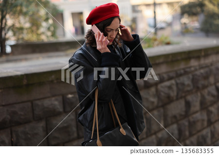 Young woman engages in a phone call while walking along a city street in a stylish beret and warm coat during autumn Young woman engages in a phone call while walking along a city street in a stylish beret and warm coat during autumn 135683355