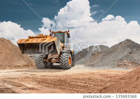 Construction site shows a wheel loader transporting sand in a bucket. The loader operates among piles of earthy materials against a cloudy sky Construction site shows a wheel loader transporting sand in a bucket. The loader operates among piles of earthy materials against a cloudy sky 135683368