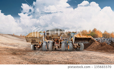 Construction equipment works on moving dirt and materials at a site in a rural area under a cloudy sky during the day. Three powerful loaders Construction equipment works on moving dirt and materials at a site in a rural area under a cloudy sky during the day. Three powerful loaders 135683370