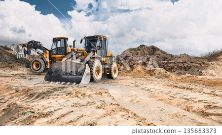 Two powerful wheel loaders stand ready to work in a sand quarry, while clouds disperse across the blue sky. The equipment is ready for construction work 135683373