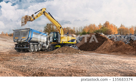 Construction workers use a loader and excavator to move dirt and materials at a site with trees and rocks in the background during daytime 135683374