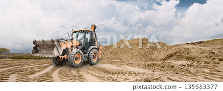 A general-purpose excavator loader moves sand at a construction site; deep track marks are visible in the ground on a cloudy day. A sand pile is visible in the background 135683377