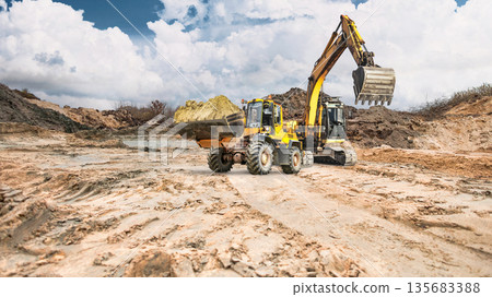 A construction loader moves soil, while in the background an excavator with its bucket raised. Clouds are visible in the sky, creating a contrast with the daytime landscape A construction loader moves soil, while in the background an excavator with its bucket raised. Clouds are visible in the sky, creating a contrast with the daytime landscape 135683388