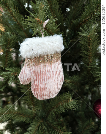 A small pink mitten made of fabric is hanging on the branches of a Christmas tree. The ornament adds a festive touch to the holiday setting 135683394