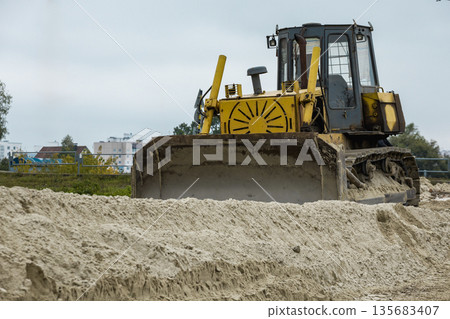 A bulldozer moves earth on a construction site in a city. The machine pushes sand in preparation for building work. Weather is cloudy and overcast 135683407